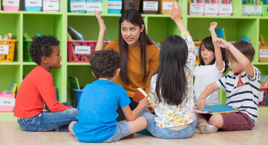 Teacher sitting on floor with students raising their hands
