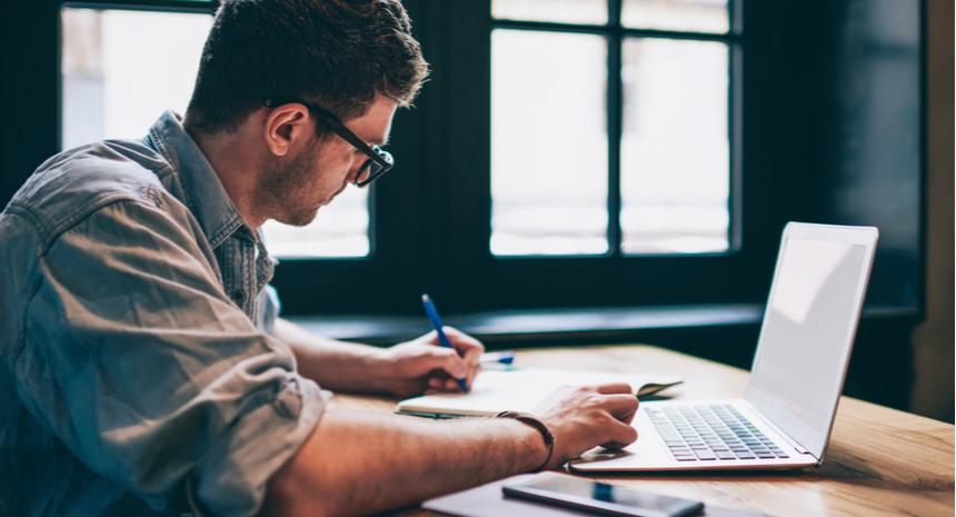 man looks a laptop and writes in notebook